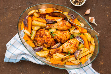 Chicken legs and thighs baked with sweet potatoes, white and purple onions, plain potatoes in a transparent baking dish on a brown concrete background. Sweet potato recipes.