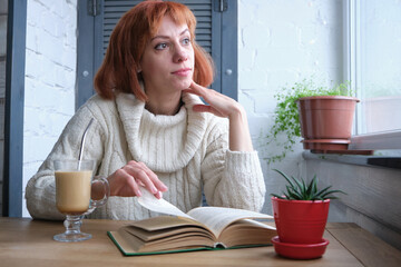 Young adult red-haired girl relaxing at home with mug of hot beverage and book in domestic kitchen, relaxing during quarantine