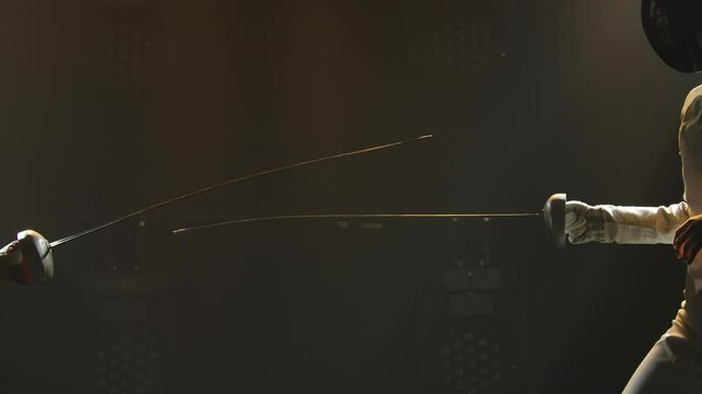 Duel of two female fencers. Professional fencers rivals demonstrate mastery of foil fencing. Black smoky background with backlit lights. Slow motion. Close up.