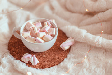 Cup of tasty cocoa drink or hot chocolate with marshmallows in cup on cork stand on a white bedspread. Winter dessert beverage. Decoration garlands of lights.