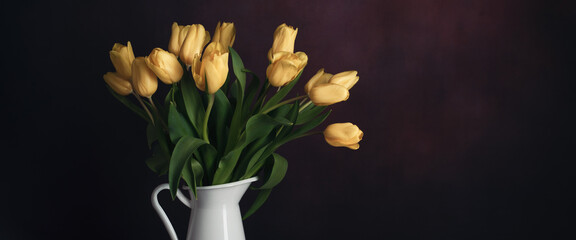 Tulips in a jug. Classic still life with a bouquet of yellow tulip flowers in a vintage white jug on a dark background and an old wooden table.