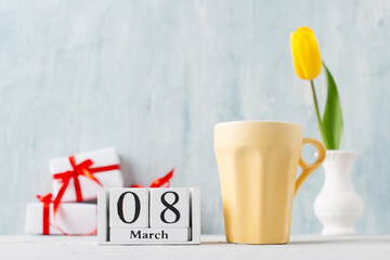 Morning coffee cup, yellow tulip in a vase, gifts and calendar March 8 on the table, on a blue background.