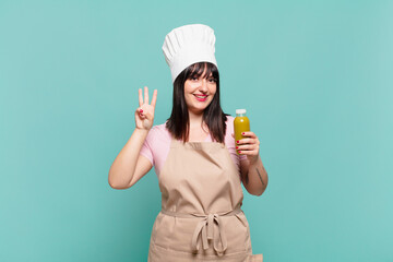 young chef woman smiling and looking friendly, showing number three or third with hand forward,...