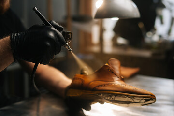 Close-up hands of unrecognizable shoemaker wearing black gloves spraying paint of light brown leather shoes. Concept of cobbler artisan repairing and restoration work in shoe repair shop.