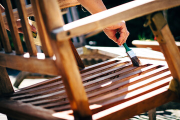 Female hand holding a brush applying varnish paint on a wooden garden chair- painting and caring for wood - obrazy, fototapety, plakaty
