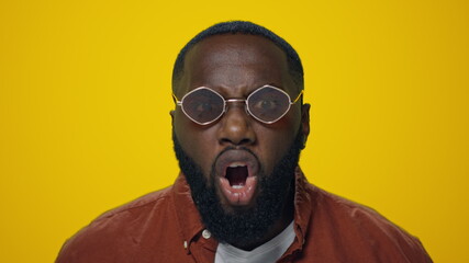 Portrait of angry african man screaming in sunglasses on yellow background.
