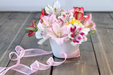 Bouquet from pink, yellow, magenta, white and red alstroemerias in white vase with pink ribbon on wooden background.