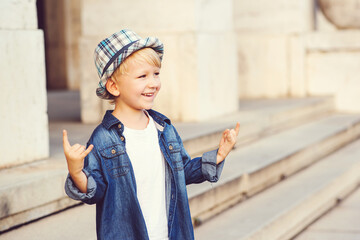 Little boy depicting heavy metal rock sign outdoors. Childhood lifestyle concept. Kid wearing denim jacket and hat.