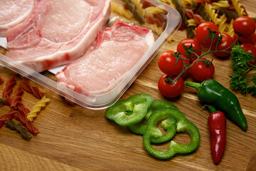 Uncooked pork steaks arranged with, fresh vine tomatoes, pepper, chilies and pasta on a wooden board