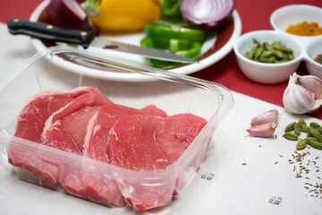 Raw beef steaks in a clear container with ingredients of red onion, green and yellow pepper, garlic, cardamom pods and cumin seeds 