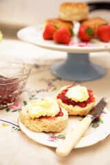 Scones with jam and cream on a plate,  with a cake stand with scones and strawberries in the background
