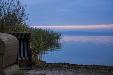 Zwei Strandkörbe  stehen verlassen an einem bewachsenen Ufer.  Es herrscht eine abendliche...