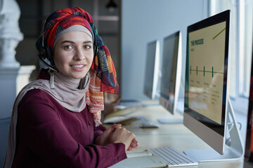 Portrait of young muslim woman in headphones smiling at camera while sitting at the table in front of computer monitor