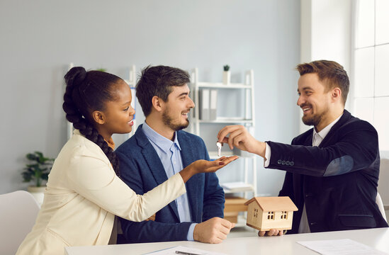 Property purchase and ownership concept. Millennial multiethnic couple taking key from new apartment buying first home together. Happy smiling realtor agent and interracial family buyers in office