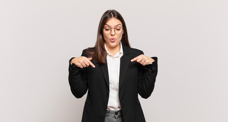 young business woman feeling shocked, open-mouthed and amazed, looking and pointing downwards in disbelief and surprise