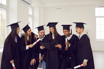 Group of students of different nationalities in graduation gowns look at photos in the graduation album in the university classroom. Concept of education, graduation, people, memories and emotions.
