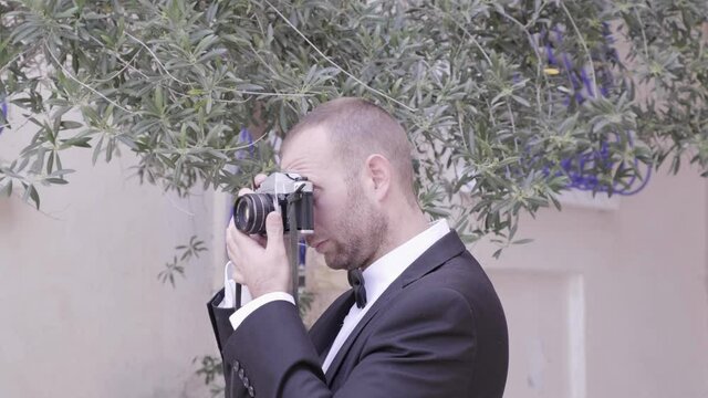 man wearing tuxedo and taking a picture with a camera. handsome white gentleman in a black suit looking around and photographing. charming guy undercover spying in a party. James bond style.