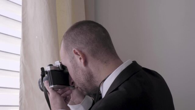 man in tuxedo taking a picture from the window. handsome guy in a black suit standing in a window with shutters and photographing. undercover James bond style spying and taking a photos.