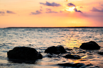 Fototapeta na wymiar Colorful sunset in Islamorada, Florida Keys with pink sky, sun behind clouds, yellow orange water waves reflection, closeup of rocks at gulf of Mexico