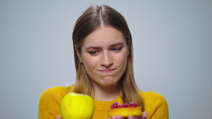 Portrait of pensive woman selecting between apple or cake on grey background.