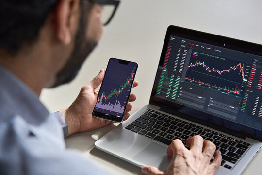 Business Man Trader Investor Analyst Using Mobile Phone App Analytics For Cryptocurrency Financial Market Analysis, Trading Data Index Chart Graph On Smartphone And Laptop Screen. Over Shoulder View