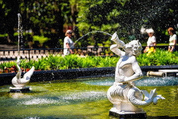 Water fountain in Forsyth park, Georgia with sculpture statues of white marble in Savannah, Georgia on sunny summer day with people in blurry blurred background - obrazy, fototapety, plakaty