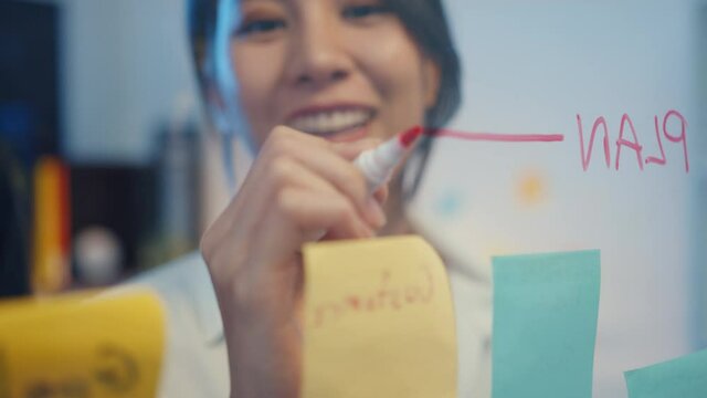 Asia businesspeople stand behind transparent glass wall listen manager pointing progress work and brainstorm meeting and worker post sticky note on wall., Business inspiration, Share business ideas.