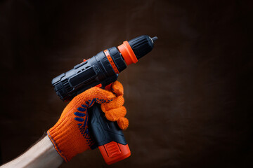 Male gloved hand holding an electric screwdriver on a dark brown background.