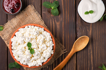 Natural cottage cheese in a traditional clay bowl, next to a wooden spoon, cranberry jam and sour cream in bowls, dark wooden background, top view. Soft curd natural healthy food, wholesome diet food