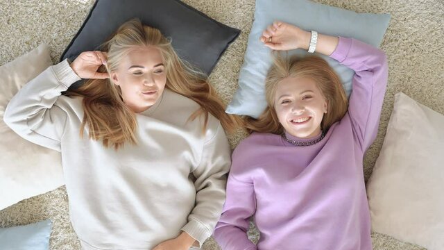 Two best girlfriends lying on the bedroom floor among pillows, relaxing, speaking and enjoying time together