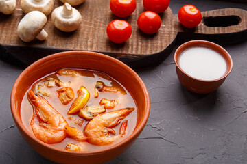 Tom yam soup with shrimps in a tureen on a concrete background next to a bowl with coconut milk mushrooms and tomatoes.