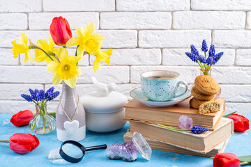 Obraz na płótnie Canvas Bouquets of spring flowers, coffee cup, books, cookies, gemstones crystal minerals on blue table on white brick background. Reading and breakfast. Concept spring, gems, hygge and cozy home interior
