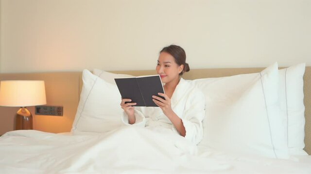 Smiling pretty Asian woman lying on the hotel white bed wearing a white dressing gown and reading a book at night time