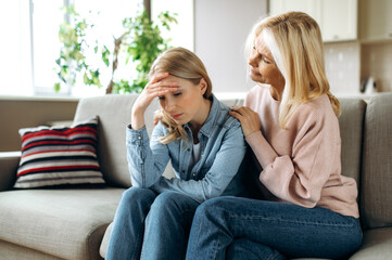 Naklejka na ściany i meble Mother and daughter together at living room. Loving mature mom sits on the couch and hugs her young adult daughter, stroking her, sympathizes, family relationship and values concept