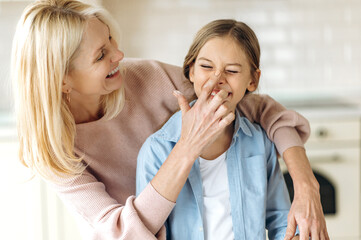 Obraz na płótnie Canvas Beautiful caucasian middle aged blonde woman and preschooler girl in the kitchen. Grandma hugging granddaughter, touching her nose finger with flour, they have fun together at home