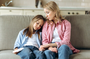 The woman and little girl are sitting at home on the couch. A young blonde mom in sits next to her...