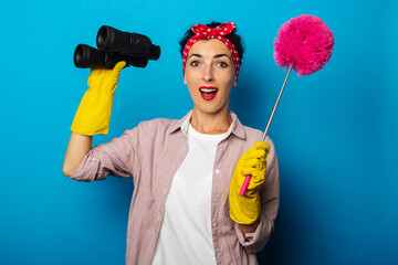 Surprised young woman in cleaning gloves holding binoculars and dust brush on blue background.