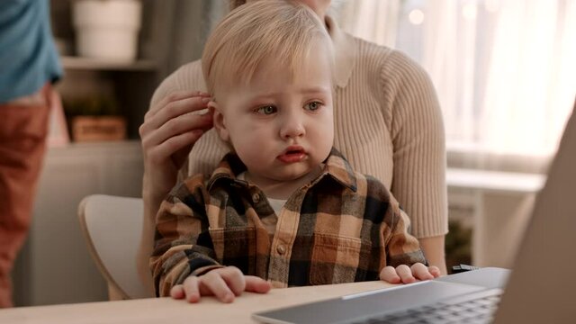 Medium close-up of adorable tear-stained Caucasian toddler wearing plaid shirt, wiping eye with hand, sitting in arms of cropped unrecognizable parent, watching laptop computer