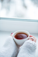 Tea drinking. The woman in a knitted, white pullover holds a cup of hot tea in her hands. Cozy weekends, winter drinks.