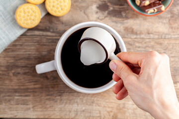 a tea time or coffee time concept with sandwich cookies, bars of chocolate aesthetic ceramic mug and plates as well as a mini creamer pitcher on wooden table. A woman is adding creamer into drink