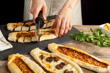 Kasarli sucuklu pide and kiymali pide are traditional Turkish flatbreads similar to pizza with meat and cheese toppings. They are served with lemon and parsley leaves. A woman is slicing them.