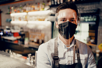 Fototapeta na wymiar A male barista employee brews and sells coffee to take away. Paper cup with a drink Americano, espresso, cappuccino. Protective mask on the face from the virus, pandemic, flu.