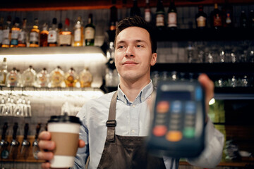 mobile phone payment NFS bank terminal. A satisfied barista brews and sells coffee to take away. Paper cup with a drink Americano, espresso, cappuccino.
