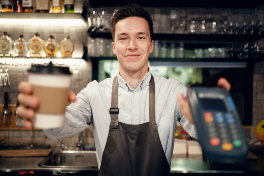 Payment via the bank's nfs terminal. Modern uniform apron in the restaurant. A satisfied barista brews and sells coffee to take away. Paper cup with a drink Americano, espresso, cappuccino.