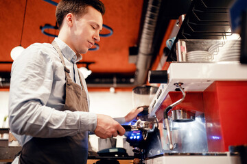 A male waiter makes coffee with a takeaway. Clothing apron uniform of the bar staff. Works in his small business restaurant cafe. Copy space. Modern interior.