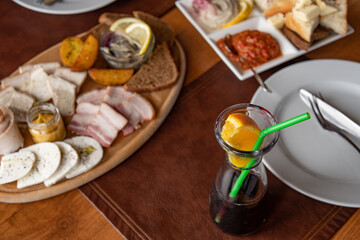 Fototapeta na wymiar Glass bottle of mulled wine with green plastic drinking straw and slice of orange citrus fruit. Top view table settings with set of appetizers on wooden tray and in white plate