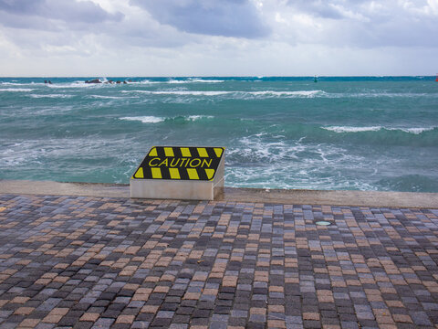 A Sign Calling For Caution Where The Waves Of The Surf Hit The Embankment As A Concept For The Uncertainty Of The Opening Of The Resort.