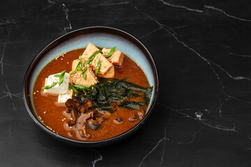 Asian miso soup with tofu, tuna, shiitake mushrooms, seaweed, sesame seeds and spring onion. Dish isolated in a blue bowl, close-up on a black marble background. Asian cuisine. Copy space.