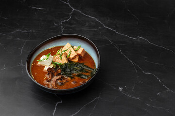 Asian miso soup with tofu, tuna, shiitake mushrooms, seaweed, sesame seeds and spring onion. Dish isolated in a blue bowl, close-up on a black marble background. Asian cuisine. Copy space.