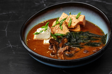 Asian miso soup with tofu, tuna, shiitake mushrooms, seaweed, sesame seeds and spring onion. Dish isolated in a blue bowl, close-up on a black marble background. Asian cuisine.